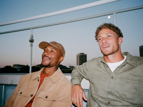 Two multiethnic male friends laughing together on a city rooftop at dusk, candid flash photo with authentic joy and urban skyline background.