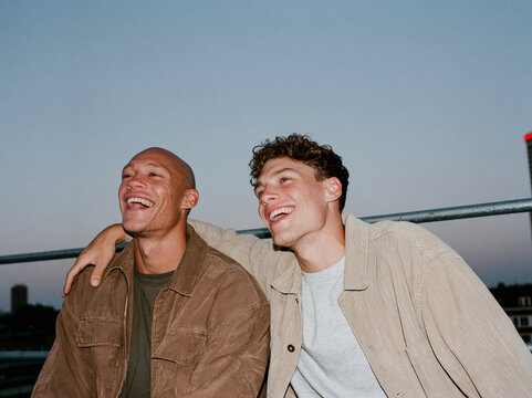 Two multiethnic male friends laughing together on a city rooftop at dusk, candid flash photo with authentic joy and urban skyline background.