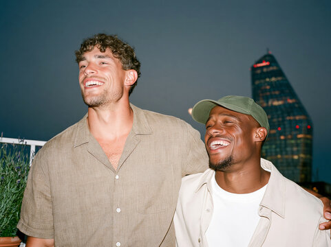 Two multiethnic male friends laughing together on a city rooftop at dusk, candid flash photo with authentic joy and urban skyline background.