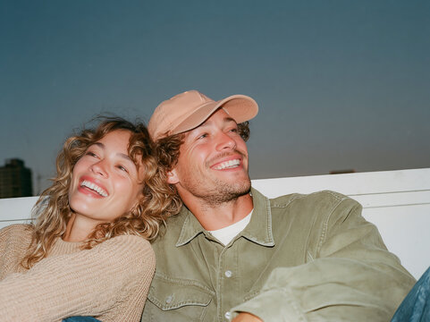 Two multiethnic male friends laughing together on a city rooftop at dusk, candid flash photo with authentic joy and urban skyline background.