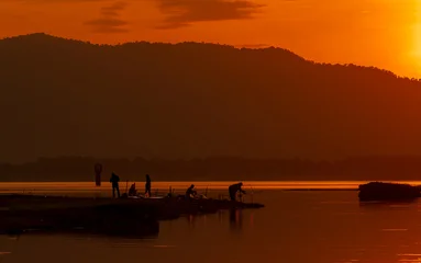 Fototapete Braun Silhouette of people fishing along a peaceful lakeshore during a golden sunrise with mountain backdrop. Tranquil nature landscape for travel, recreation, outdoor lifestyle and tourism concepts.  © Fahroni