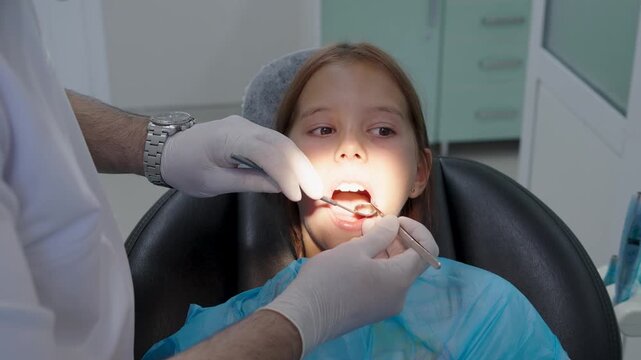 Pediatric dentist examining teeth of a brave girl sitting in dental chair.