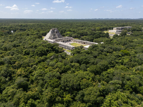 Aerial view of the ancient Maya ruins of Uxmal, featuring the Pyramid of the Magician surrounded by dense green jungle under a blue sky in Uxmal, Yucatan, Mexico.