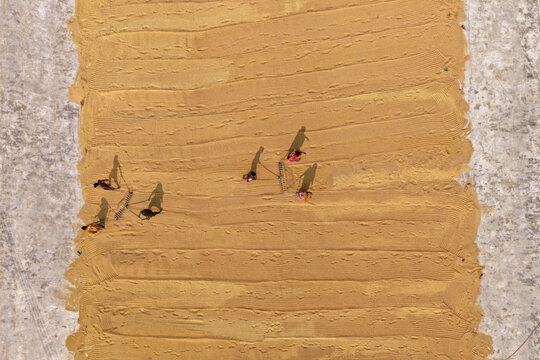 Aerial view of workers using rakes to spread golden rice grains across a large drying field in Ashuganj Upazila, Chittagong Division, Bangladesh.