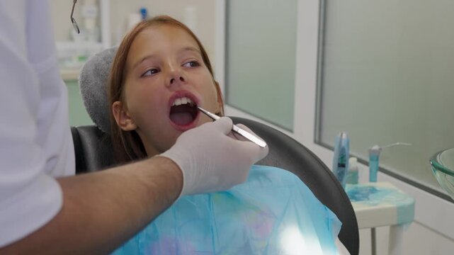 Pediatric dentist examining teeth of a brave girl sitting in dental chair.