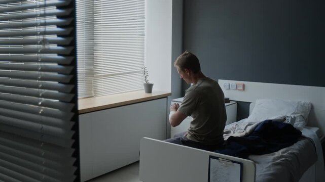Young adult Caucasian male patient with bandaged arm sitting on hospital bed, adjusting dressing and looking out window. Ideal for medical insurance and patient recovery campaigns