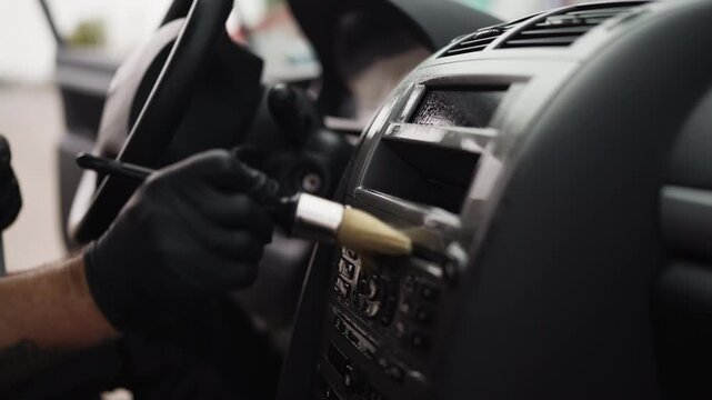 Professional auto detailer wearing protective gloves using a special soft brush to remove dust from the dashboard, radio controls, and air conditioning vents inside a modern vehicle