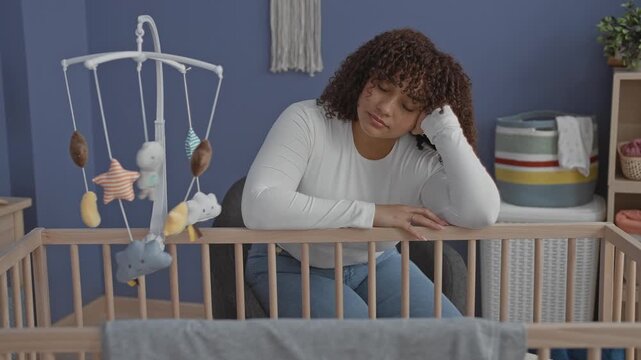 Woman leaning on crib with hand to head, touching mobile and peering into empty cradle in building; exhaustion.