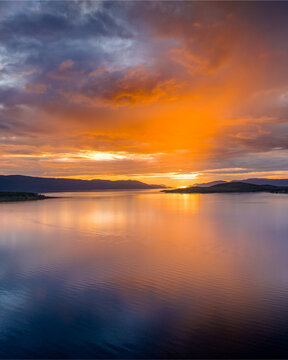 Aerial view of Malangen fjord at sunset with calm reflective water and distant mountains under a vibrant orange and purple sky in Troms, Norway.