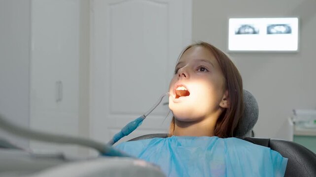 Pediatric dentist examining teeth of young girl sitting in dental chair.