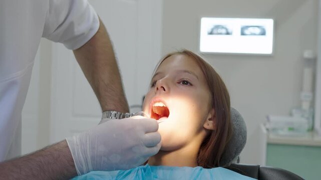 Pediatric dentist examining teeth of young girl sitting in dental chair.