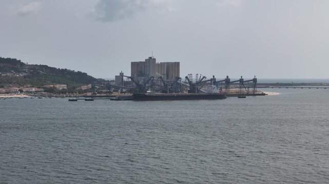 Trafaria, Setubal, Portugal - 25 July 2026: Aerial view of the Silopor grain terminal with a large cargo ship docked at the pier, industrial cranes, and coastal landscape.