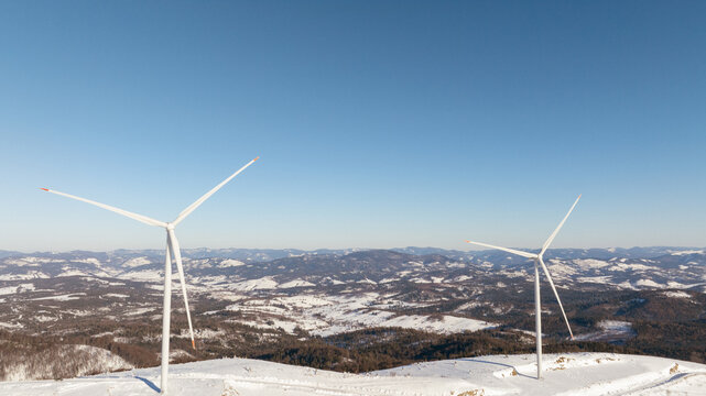 Wind turbines stand tall on a snow-covered mountain range under a clear blue sky