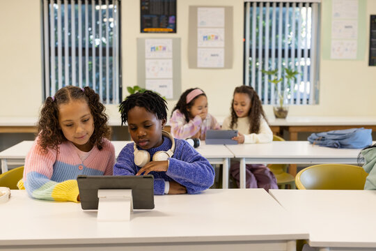 Diverse youth females sharing tablet on stand at front row in classroom, using headphones