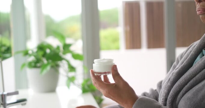 Senior African American woman in robe opening jar, applying morning cream at vanity, copy space