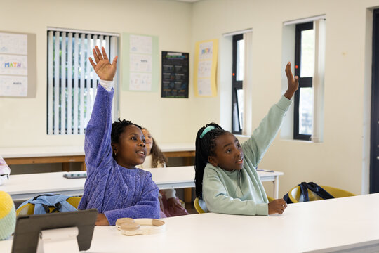African American girls raising hands while sitting at classroom table with tablet and headphones
