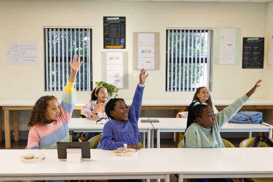Female school-age students raising hands at classroom tables with tablet stand and headphones