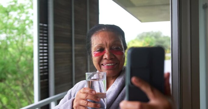 Senior African American woman wearing robe and eye patches facing phone, sipping water on balcony