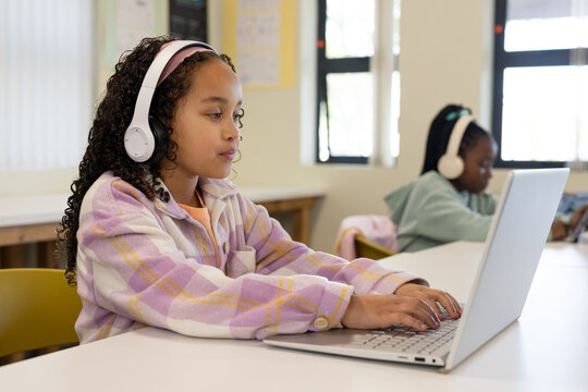 Two female school-age students typing in class on laptops, left wearing plaid-fleece and headsets