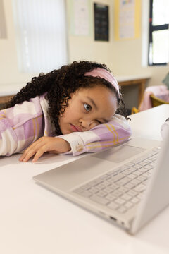 African American preteen girl resting at school desk, gazing at silver laptop wearing pink headband