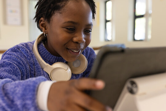 African female child leaning forward using tablet in classroom, wearing blue sweater and headphones