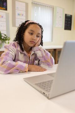 School-age student sitting at table in classroom in pastel jacket and pink headband using laptop