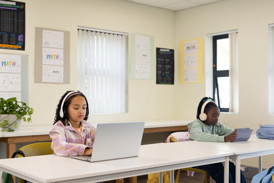 African American teen females typing on laptop and tablet at classroom desks wearing white headsets