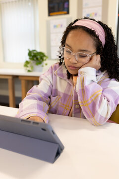 African American girl wearing pastel plaid jacket studying at classroom table using tablet