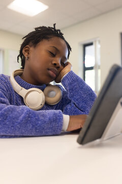 African American child napping at school desk, wearing blue sweater and headphones, tablet nearby