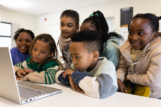 Diverse children gathering around silver laptop on table in classroom, wearing sweaters