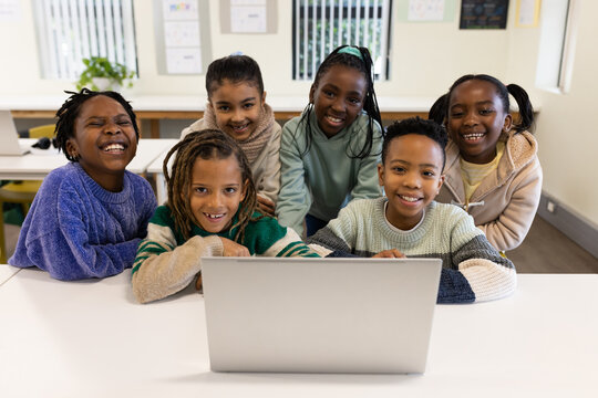Diverse students leaning and smiling around silver laptop on white table in classroom with posters