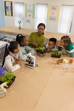 African American teacher pointing, teaching 5 kids at table with scope and seedling trays in lab
