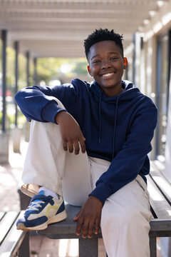 Teenage male smiling, sitting on wood bench under covered path wearing navy hoodie and beige pants