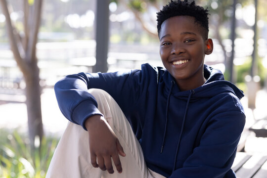 African American teenage boy sitting on wooden bench at sunlit park wearing navy hoodie, smiling