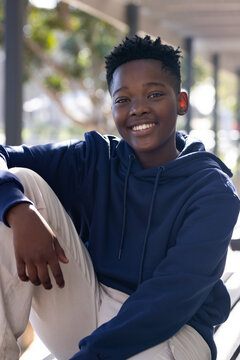 African male youth sitting on bench under pergola wearing navy hoodie and light pants smiling