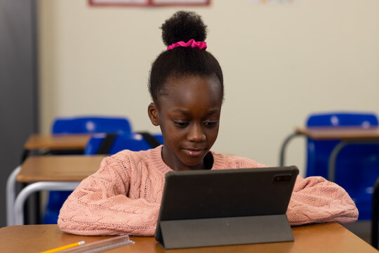 African American child wearing pink sweater scrunchie at school desk using tablet in folio, pencil