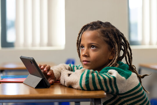 African American boy sitting at desk in class, wearing green striped sweater, using tablet folio