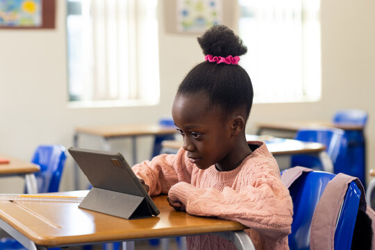 African American child wearing pink sweater sitting at classroom desk, focusing on tablet in folio