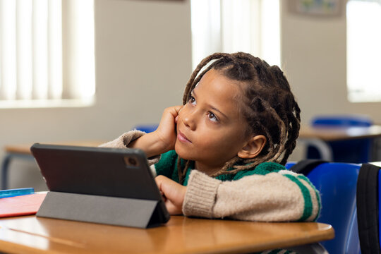 School-age African American boy sitting at desk in classroom, gazing at tablet and red notebook