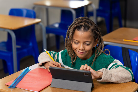 African American child sitting at school desk using tablet in folio with pencil, in green sweater