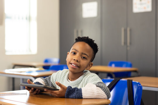 African American schoolboy holding tablet, sitting at desk in classroom with blue chairs