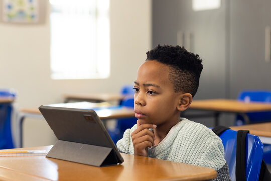 African American preteen student boy studying at wooden desk in classroom, using tablet and pencil