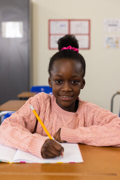 African girl child sitting at wooden desk in classroom holding yellow pencil over open notebook