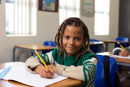 School-age African American boy sitting at classroom desk holding yellow pencil writing in notebook
