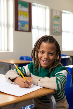 African American schoolboy writing with yellow pencil on paper at wooden desk wearing green sweater