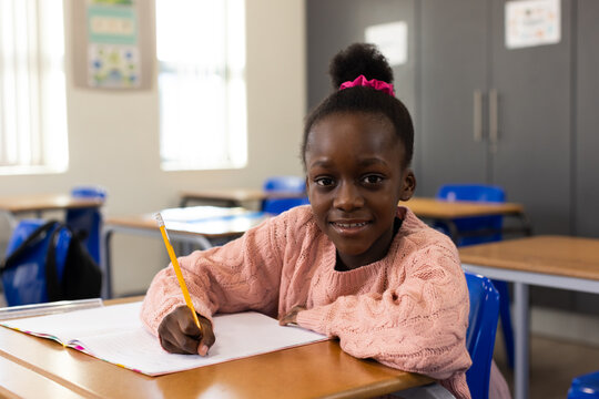 African child girl sitting at school desk in pink sweater holding yellow pencil on lined notebook