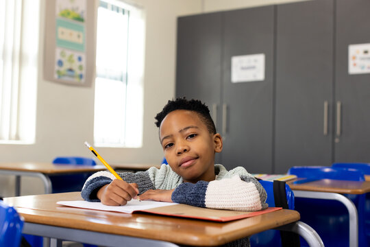 African youth male student writing in notebook at wooden desk holding yellow pencil wearing sweater