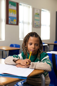 African American boy sitting in class in sweater holding yellow pencil near mouth notebook on desk