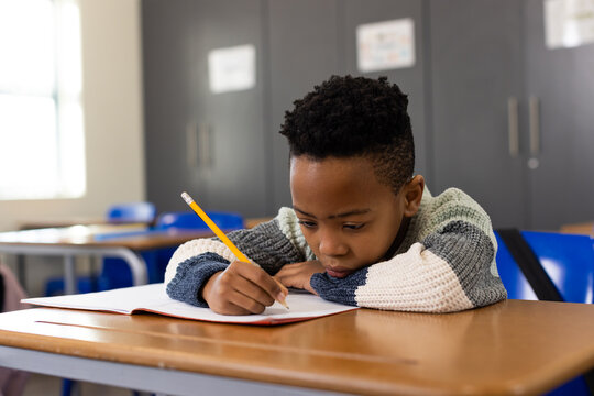 Male child wearing color-block sweater, writing in notebook at classroom desk with yellow pencil