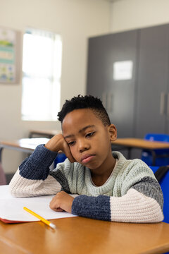 African American boy sitting at desk in classroom, knit sweater, gazing at paper with yellow pencil